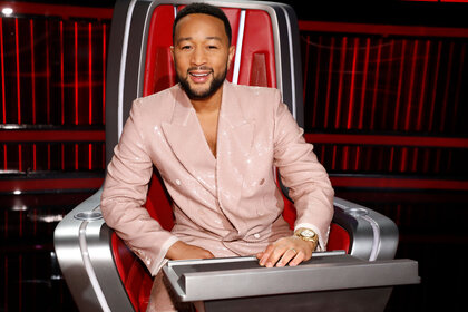 John Legend posed and smiling while sitting in his chair on the set of The Voice