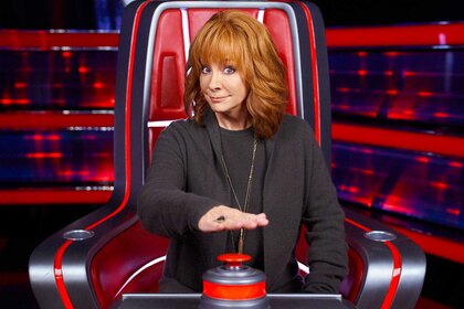 Reba McEntire in the middle of pushing a button while sitting in a judges chair for The Voice.