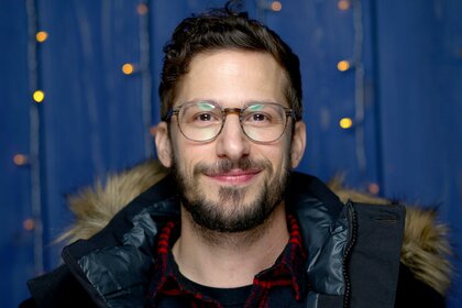 Andy Samberg, wearing a black coat with glasses on, smiles at the camera.