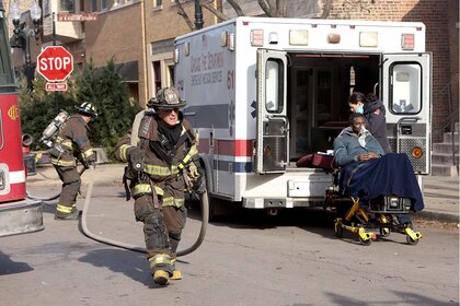 Christopher Herrmann holds a fire hose while Violet Mikami tends to a Coughing Victim in a gurney near an ambulance in Chicago Fire Episode 1202.