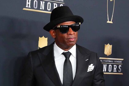 Deion Sanders wears a black hat, sunglasses, and suit.