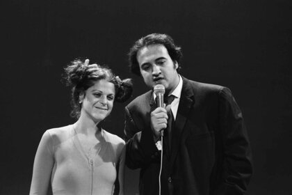 Gilda Radner smiles next to John Belushi who speaks into a mic on Saturday Night Live Season 1 Episode 23.