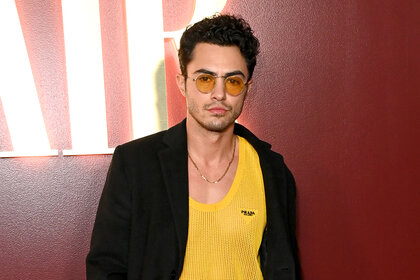 Darren Barnet wears a black blazer and yellow shirt and sunglasses in front of a red wall