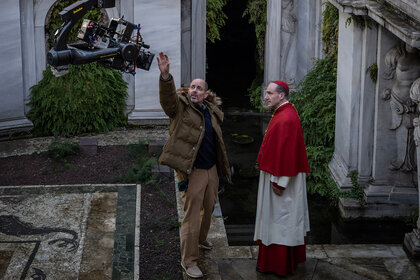 Director Edward Berger and Ralph Fiennes point to a statue behind the scenes of Conclave