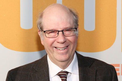 Stephen Tobolowsky smiles wearing a brown suit and glasses on the red carpet