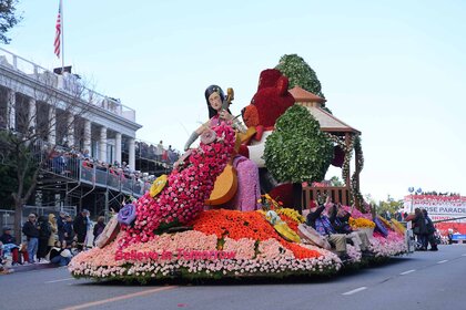 A floral arrangement parade float in the shape of a woman playing a cello is walked on a road.