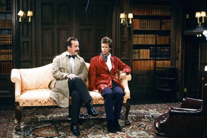 Inspector Langley (Phil Hartman) and Sir Jeffrey Kurr (Dana Carvey) sit on a sofa together during an SNL sketch.