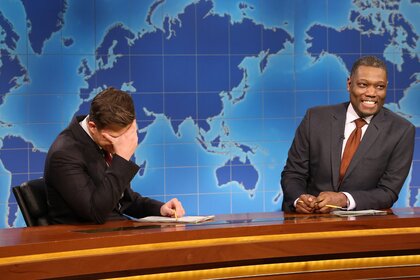 Colin Jost and Michael Che laughing while seated behind a desk.