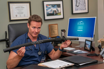 Dr. Bruce holds up a sword at his desk on St. Denis Medical Season 1 Episode 6