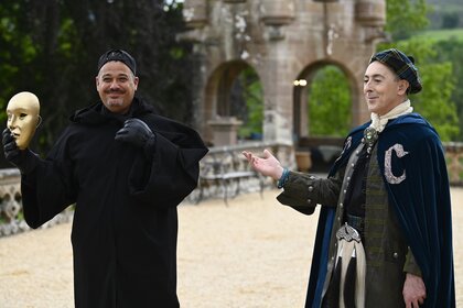 Alan Cumming gestures to Rob Mariano holding a mask while wearing a cloak on The Traitors Episode 303.