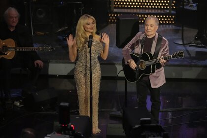 Sabrina Carpenter standing behind a microphone next to Paul Simon playing guitar.