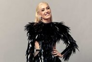 Gwen Stefani, wearing a black feather top, posing for the camera and smiling with her left arm on her hip.