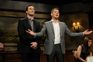 Michael Buble stands in front of Jon Hamm with his arms outstretched towards the audience