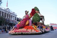 A floral arrangement parade float in the shape of a woman playing a cello is walked on a road.