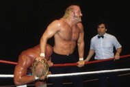 Jesse Ventura holds down Hulk Hogan's head in a wrestling ring as a referee approaches.