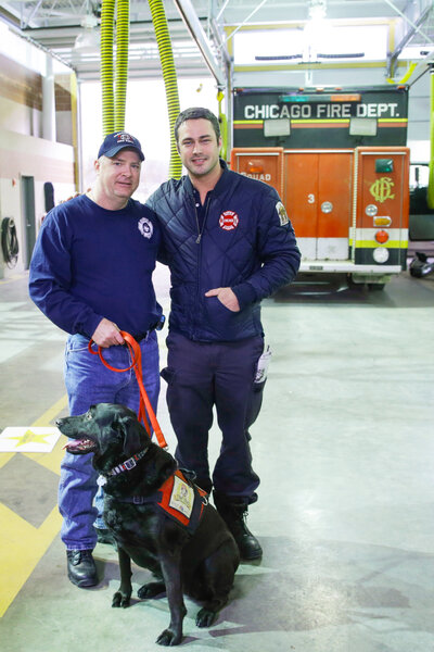 Jacksonville Fire Lt. Todd Warrick, Smokey the Labrador Retriever, and Taylor Kinney posing together in front of a firetruck.