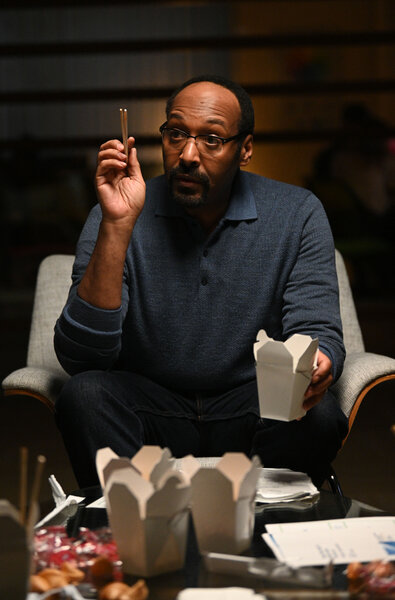 Alec Mercer sitting at a table with his hand raised and eating takeout food.