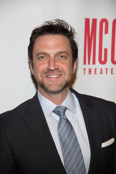 Raul Esparza smiling in a suit in front of a step and repeat.