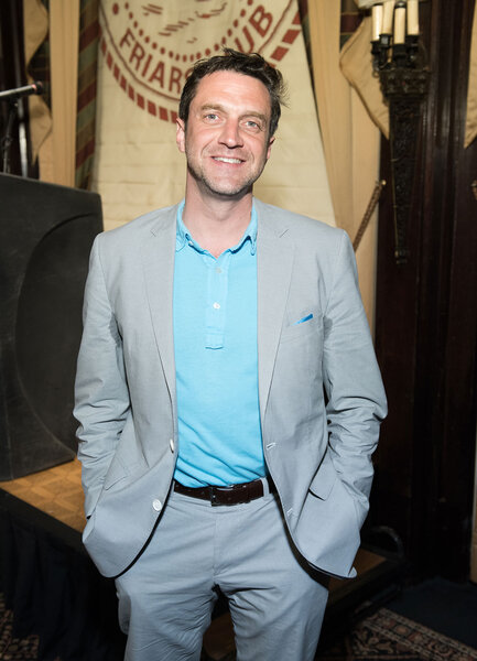 Raul Esparza standing in a suit and smiling.