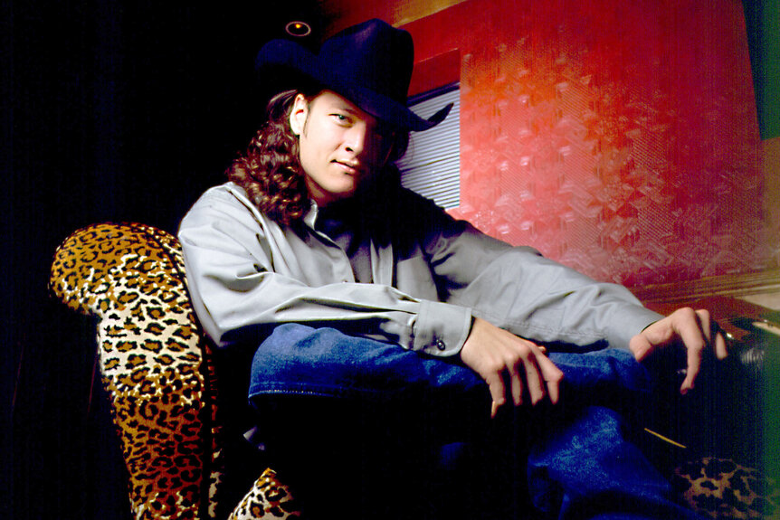 Blake Shelton sits on a leopard print chair with a cowboy hat and long curly hair