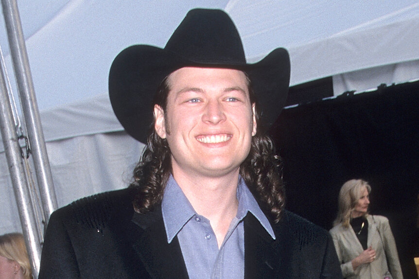 Blake Shelton smiles wearing a cowboy hat with long curly hair