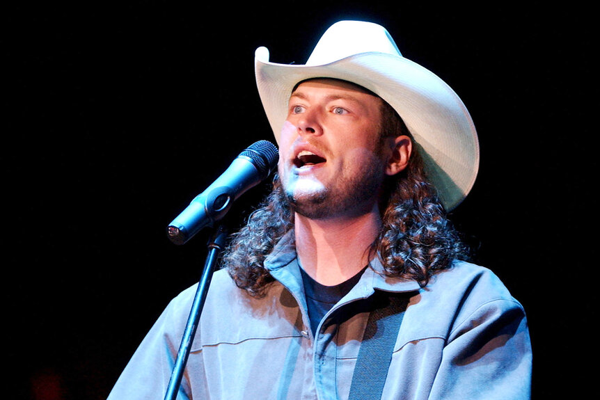 Blake Shelton sings on stage with a white cowboy hat and long curly hair