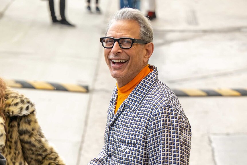 Jeff Goldblum smiling outside wearing a checkered blazer with a orange turtleneck.