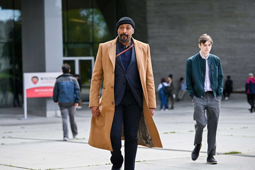 Alec Mercer appears in a tan trench coat walking on a campus.