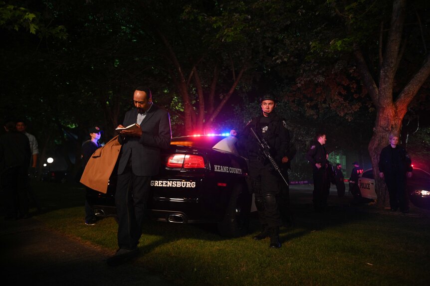 Alec Mercer leaning against a police car in the middle of an investigation.