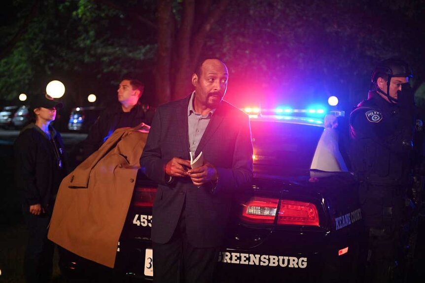 Alec Mercer leaning against a police car in the middle of an investigation.