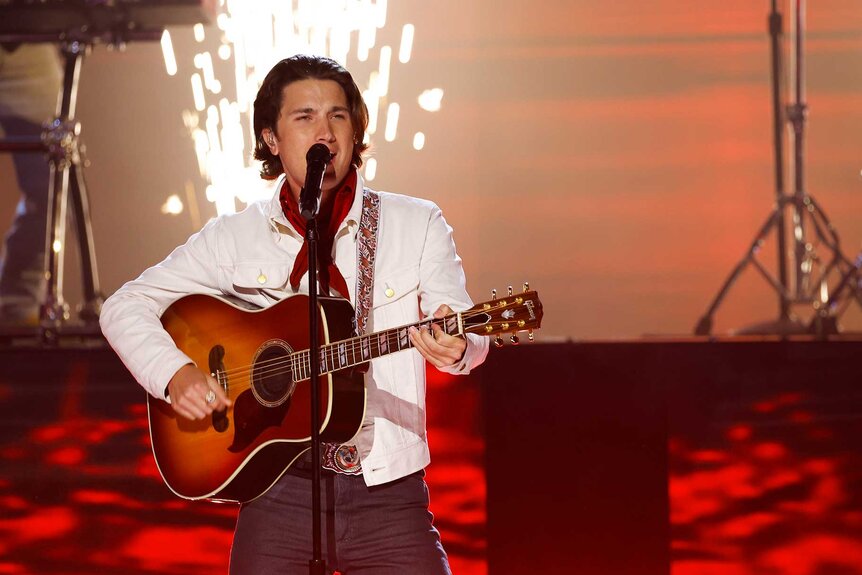 Drake Milligan playing the guitar and singing on stage while sparks appear in the background.