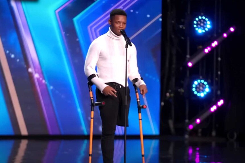 Musa Motha, wearing a white shirt with his crutches, standing in front of a mic on stage.