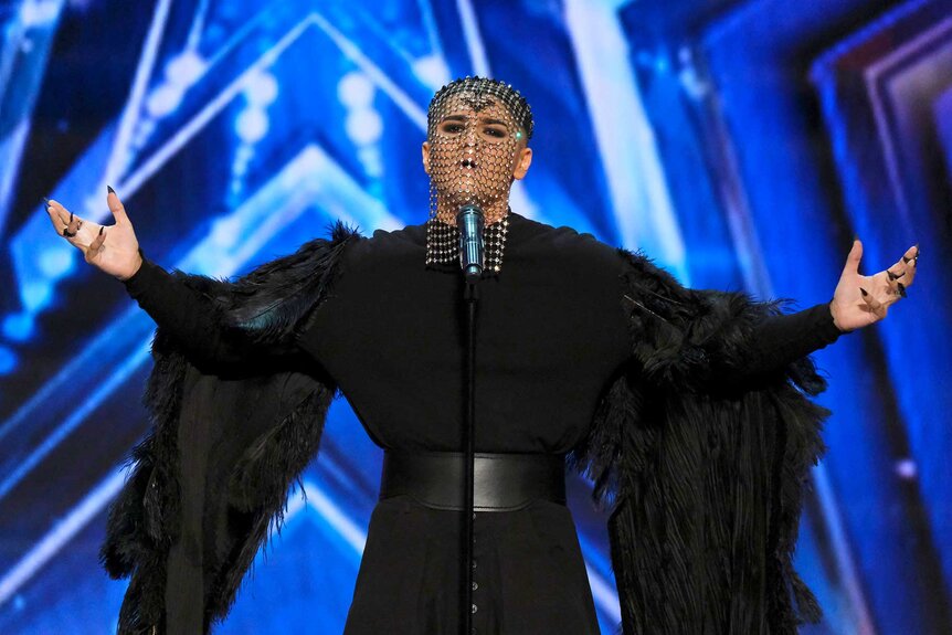 Sheldon Riley, wearing a long black dress with long sleeves with a chain mask, on stage standing in front of a mic.