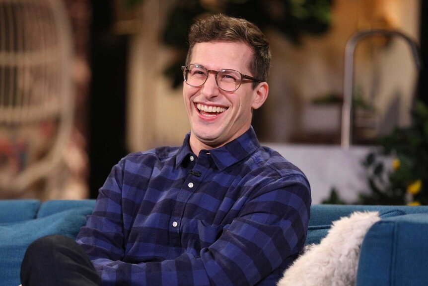 Andy Samberg, wearing a plaid purple shirt, laughing and smile at the camera while sitting on a blue couch.