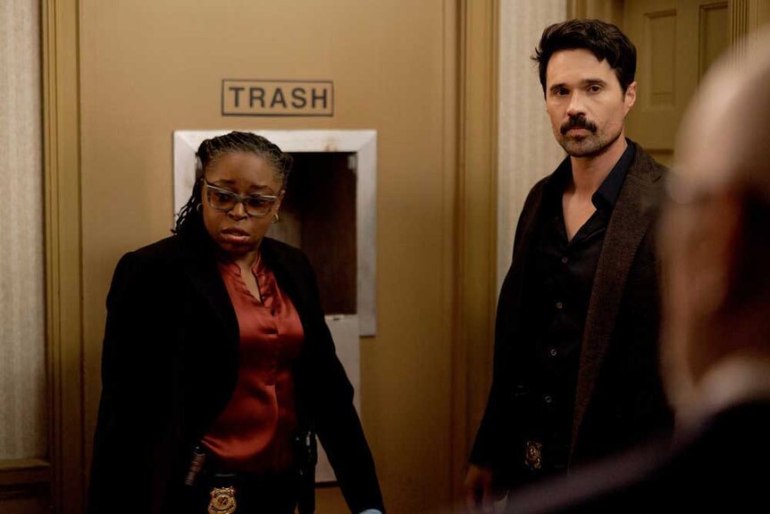 Detective Shaker standing next to Detective Mark Trent in front of a door that says "Trash."