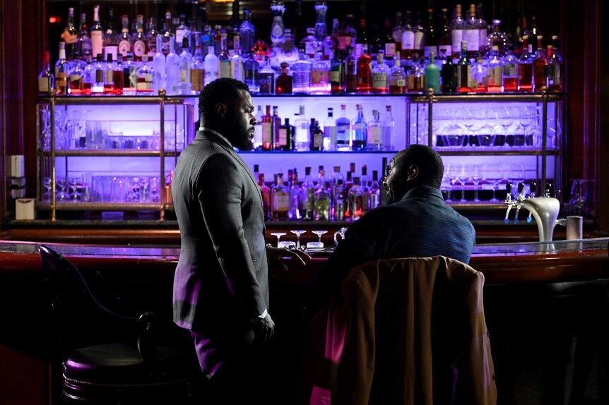 Dustin Atwood standing next to Alec Mercer, who's sitting at a bar with purple lights in the background.