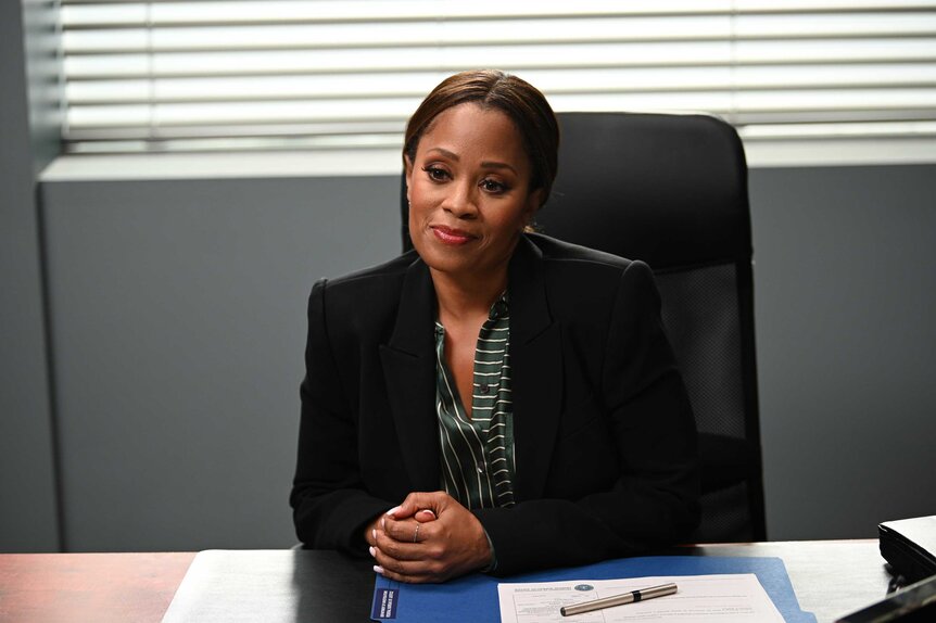 Marisa, wearing a black blazer, sitting at a desk with her hands folded together.