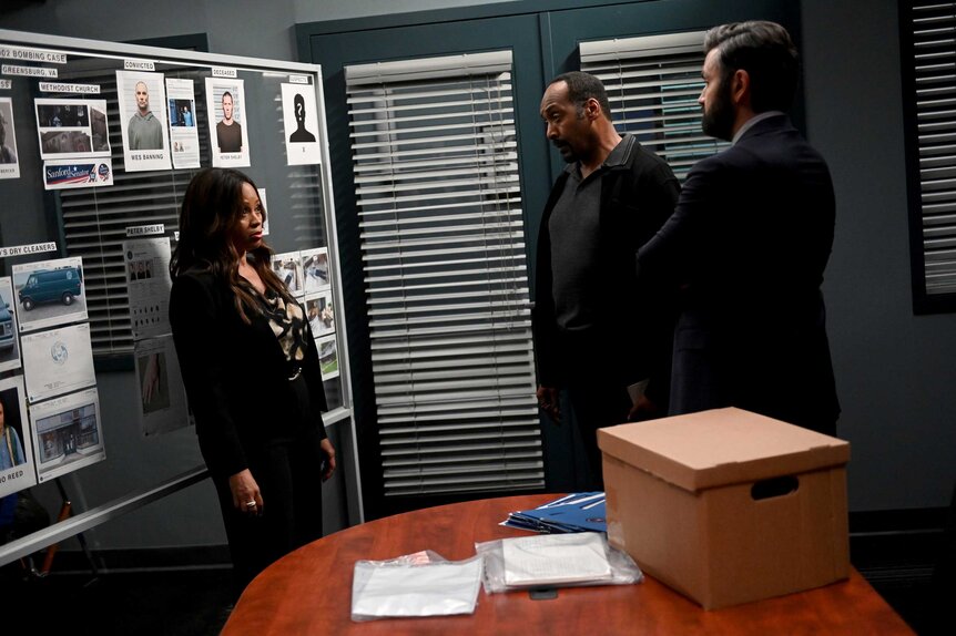 Marisa, Alec Mercer, and Jace Richards looking at each other while standing next to a wood table with a box on it.