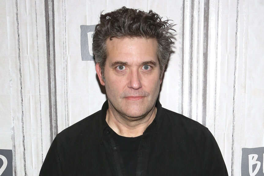 Craig Bierko stands in front of a step and repeat in a black shirt