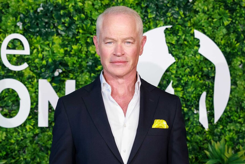 Neal McDonough, wearing a black blazer, poses in front of a green backdrop.