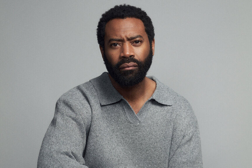 A headshot of Nicholas Pinnock wearing a grey sweater against a grey backdrop