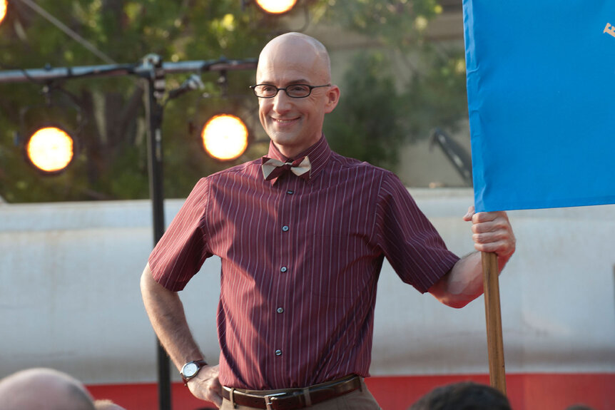 Dean Pelton (Jim Rash) smiles while holding a large flag