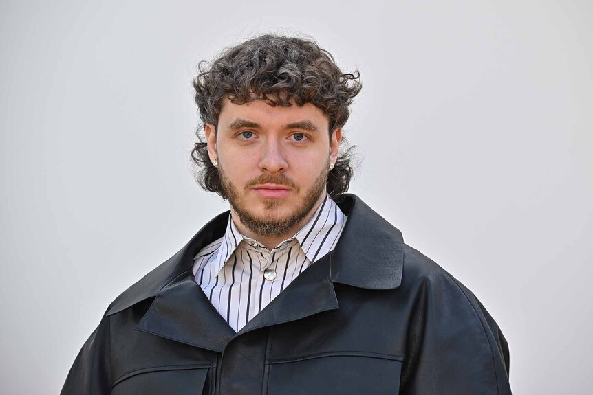 Jack Harlow smiles in a striped shirt and grey jacket.