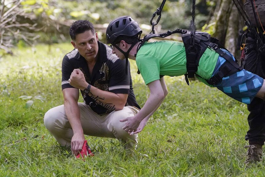 Joe Manganiello crouches next to Aron Barbell who is on a zipline on Deal or No Deal Island Episode 103