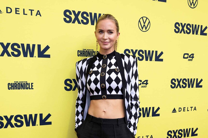 Emily Blunt smiles in a black and white checkered jacket.