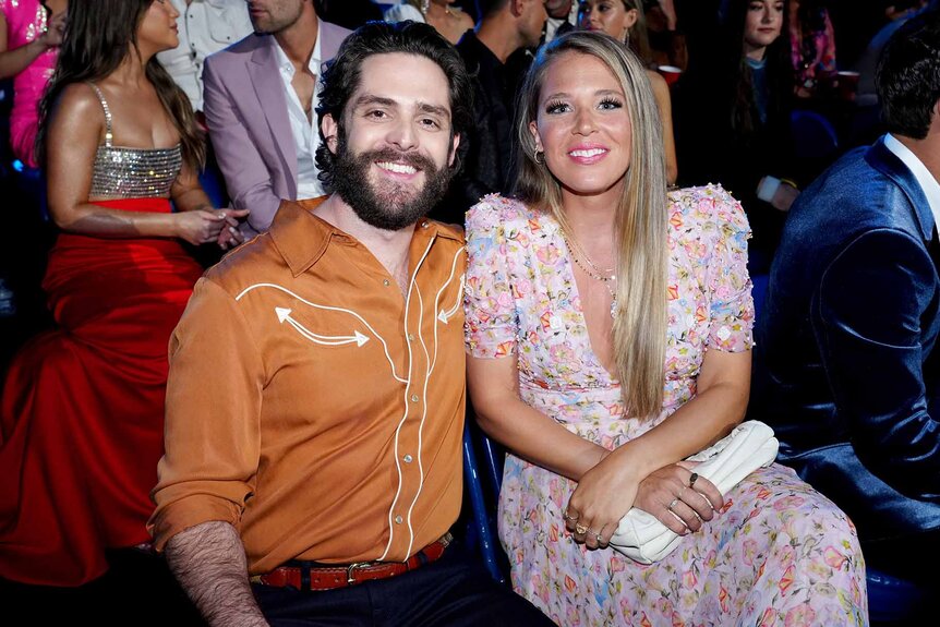 Thomas Rhett wears an orange shirt and smiles with wife Lauren Akins who wears a pink dress.