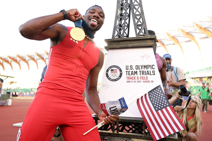 Noah Lyles holds up his medal holding an american flag in front of a mini eiffel tower