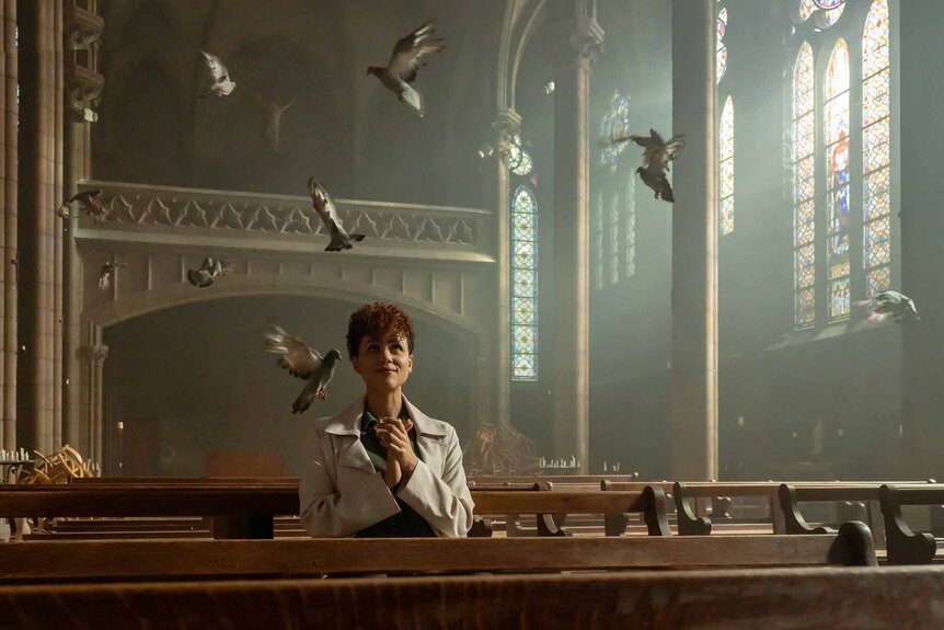 Zee (Nathalie Emmanuel) sits in church pews with birds flying above in The Killer (2024).