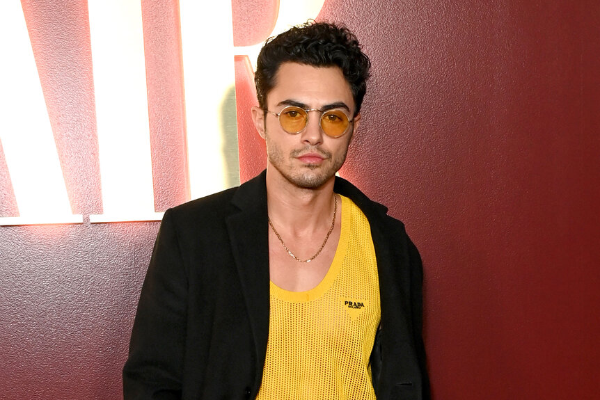 Darren Barnet wears a black blazer and yellow shirt and sunglasses in front of a red wall