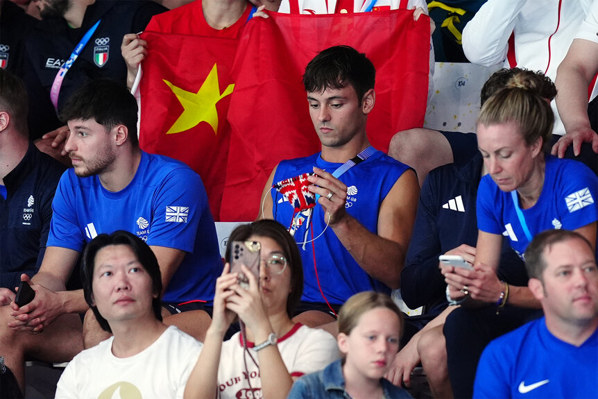 Tom Daley knits while sitting in the stands during the womens Synchronized 3m Springboard Final at the 2024 olympics